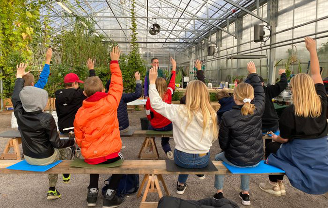 Elever som sitter i ett växthus och räcker upp händerna