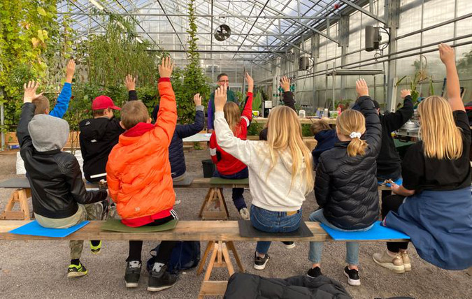 Elever sitter på bänkar i ett växthus och räcker upp händerna.